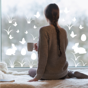Frau sitzt mit einer Kaffeetasse in der Hand vor einem großen Fenster, auf dem weiße Aufkleber in Form von Osterhasen, Ostereier und Grashalmen geklebt sind
